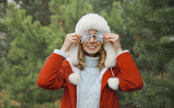 Winter day, happy smiling young woman with snowflakes decoration toys having fun enjoys snow, warm weather against Christmas tree, joyful girl in white hat standing in snowy forest