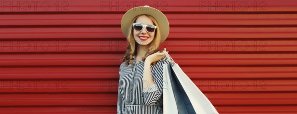 Stylish beautiful happy smiling young woman posing with shopping bags in summer straw hat on red wall background