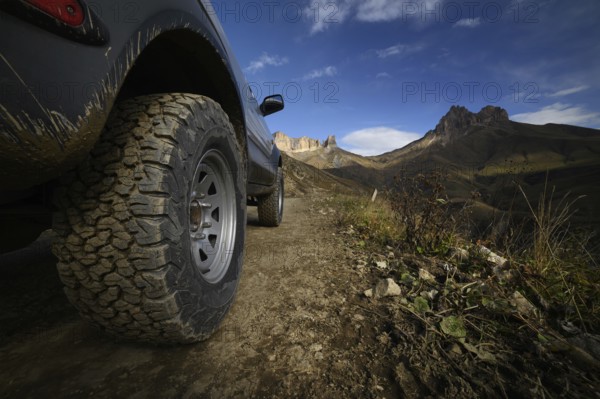 Close-up A large off-road all-terrain car wheel stands on a rough road in the mountains against the backdrop of rocks. Off-road tours and off-road travel concept