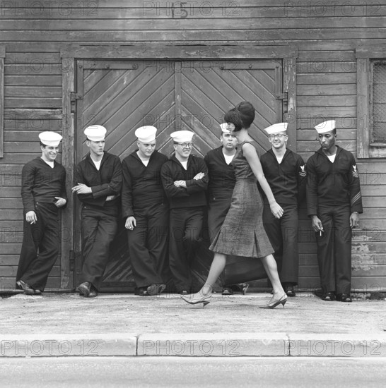 Fashion in the 1960s. A group of American sailors are standing at the docks on shore leave and makes a nice addition to the fashion shoot. A young model passes them wearing a dress with matching handbag and shoes. Sweden 1964 ref BV59-5
