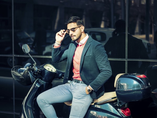Young stylish man in suit and sunglasses standing by his classic Italian scooter on empty road in sunny city