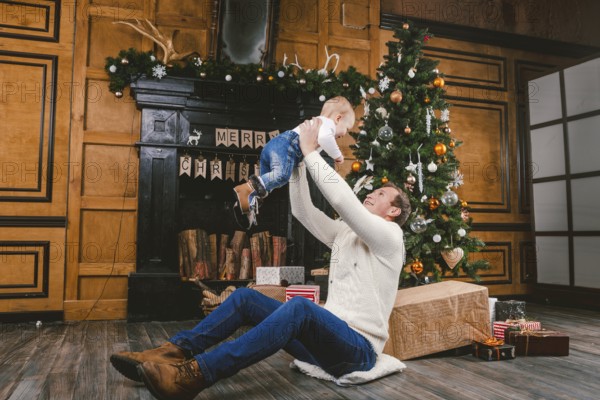 Happy father and son at home near Christmas tree. New Year and Christmas theme. Holiday mood. Family winter holidays concept. Father raises son in arms sitting on floor near Christmas tree and gift
