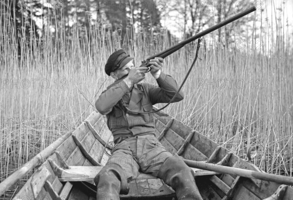 In the 1930s. A man sits in a rowing boat and aims his rifle upwards to shoot ducks. Sweden 1939. Kristoffersson ref 155-11