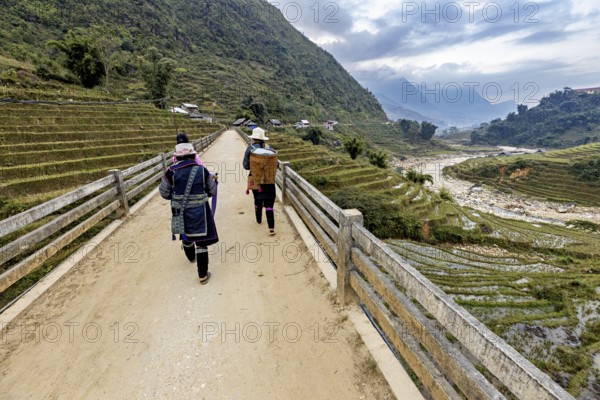 Two people walk on a bridge with a view of terraced rice paddies and mountains, local women farmers walk through the countryside near Sa Pa in Vietnam