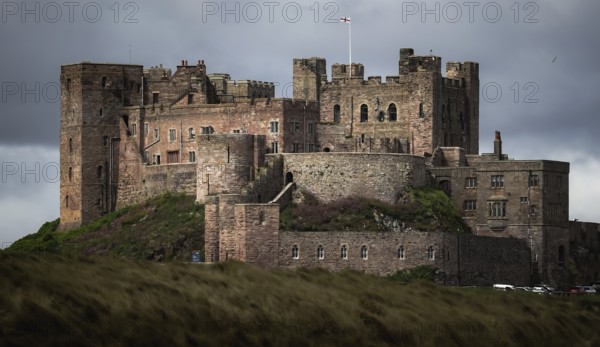 Bamburgh Castle, a medieval stone castle stands majestically under a cloudy sky, surrounded by grassy hills along the shoreline of the North Sea in Northumberland County, England, United Kingdom
