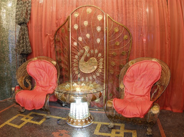 Nice metal delicate forged furniture in oriental style, peacock theme prevailing: two armchairs, glass coffee table with lighting inside its stand and folding screen against red orange curtains