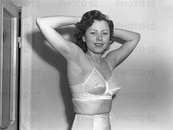 Woman of the 1940s. A young woman in a photographers studio. She holds up her arms and hands and this way covers her face partly. She is wearing a white bra and corset. Sweden 1949. Kristoffersson ref 261A-13