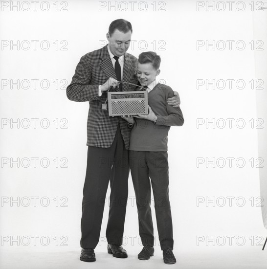 In the 1960s. Father and son with a radio. It's a typical 1960s radio with a cover of perforated leather. Battery operated transistor radio with AM and FM bands. The radiostations and frequenses are displayed on the front, and a knob to turn and adjust to get best possible reception. Sweden 1960s CX88-1