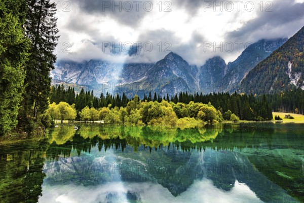 The sun's rays break through the clouds. The smooth surface of the water reflects forest and mountains. Gorgeous reflections. The lake Fusine in Northern Italy. Mountains covered in mist