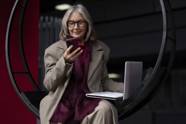 A senior businesswoman engages in work on her laptop outside, occasionally checking her smartphone. She is dressed in professional attire and appears focused while sitting in a contemporary setting