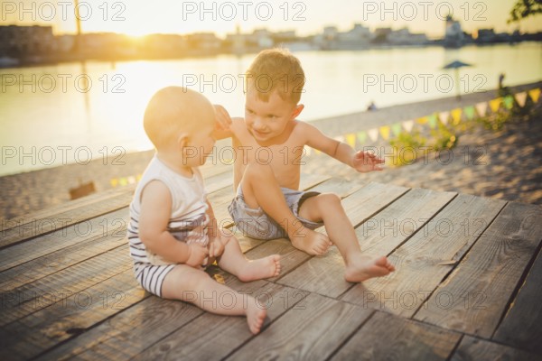 Two little boys sitting on the pier on the river bank. Concept of friendship and fraternity. Sunny joyful summer day. Children sitting on pier. Two children of different age sitting on a wooden pier