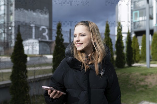 A young woman stands outdoors, smiling while using her smartphone. She is surrounded by modern architectural buildings and greenery under a cloudy sky
