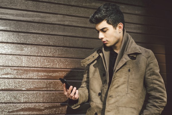 Portrait of young handsome Turkish male model of Mediterranean race brunet in leather coat using hand phone of technology businessman on street in sunny weather against background of wooden wall