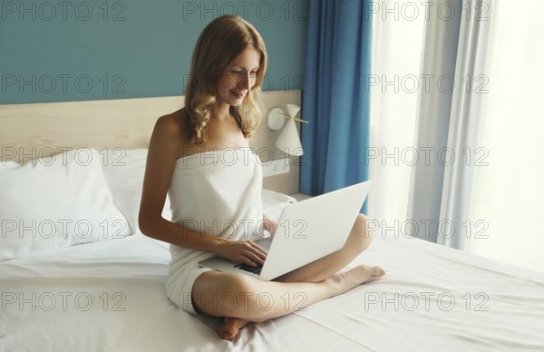 Happy young woman working on a laptop at home in the morning sitting on the bed in bedroom