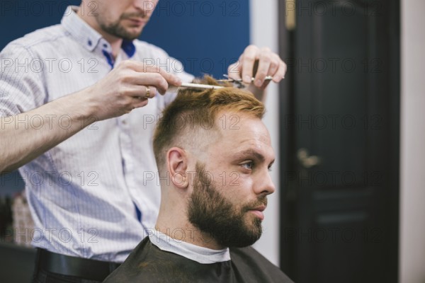 Barber Making Haircut Bearded Man In Barbershop. Professional stylist cutting client hair in salon. Barber using scissors and comb. Skillful hairdresser cutting male hair. Hair Care Service Concept