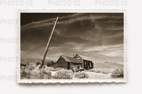 Lonely wooden house in Bodie, in front of it a crooked lamp post. Dry grass and undergrowth in the foreground. Photo in sepia tone with a white jagged edge and a sunburst shadow. Lonely wooden house in Body, besides a crooked lighting mast. In the foreground dry graß and dense undergrowth. Photo in sepia toning with white jagged edges
