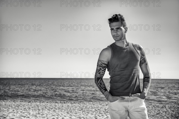 Three Quarter Shot of a Handsome Athletic Young Man in Trendy Attire, on a Beach in a Sunny Summer Day, Looking At Camera against Blue Sky Background