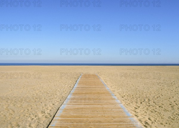 A yellow-blue picture, where the blue half is sky and the yellow half is sand and a wooden path on it. A thin dark blue line of sea in the middle