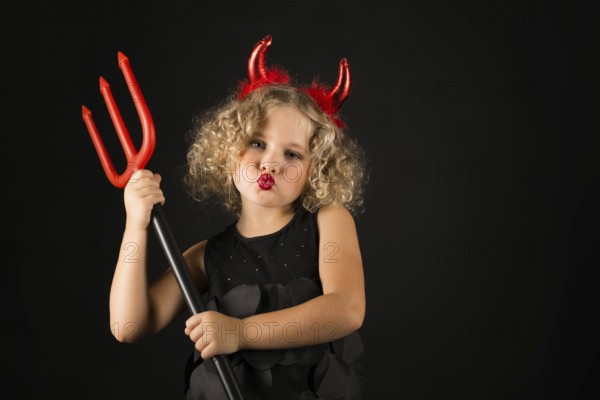 Isolated on black, adorable caucasian blonde little girl in black dress, with red horns on head hold trident both hands, look at camera, serious look