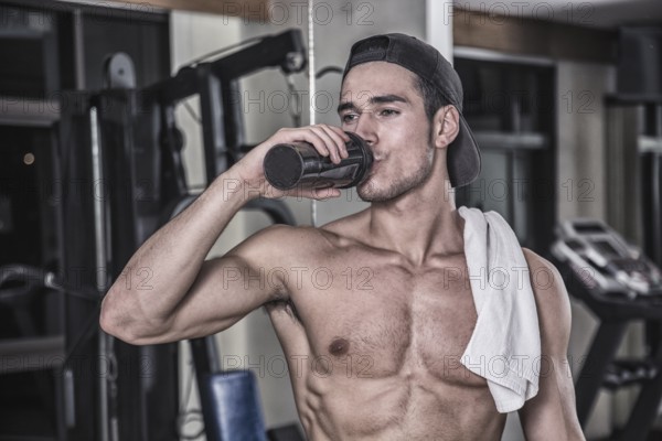 Attractive athletic shirtless young man drinking protein shake from blender in gym while looking at camera
