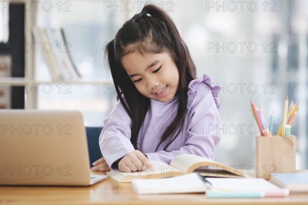 A young girl is sitting at a desk with a laptop and a book. She is smiling and she is enjoying her work