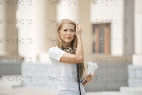 Cheerful young woman with vintage headphones around her neck, holding a coffee cup with a straw and a tablet PC, against an urban backdrop