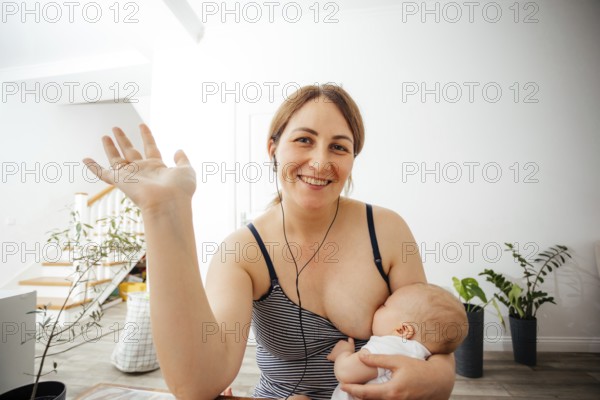 Portrait of cheerful mother in casual top nursing cute newborn while online meeting. Woman combines remote work and raising her child. Webcam view woman looking at the camera
