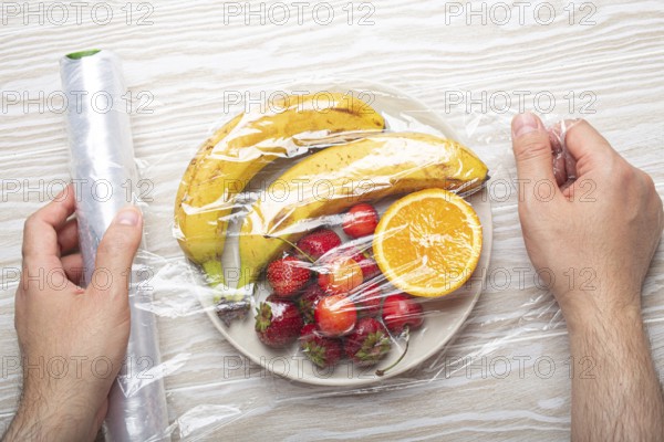 Male hands wrapping assorted different fruit and berries on white plate in plastic clear film top view on white wooden rustic table background, food photography, food photography