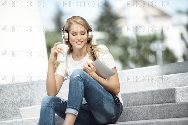 Happy young woman with vintage headphones, holding a takeaway coffee mug and a tablet PC, sitting on a staircase against an urban background