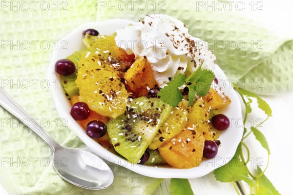Fruit salad with orange, kiwi, cranberries and baked pumpkin, whipped cream, sprinkled with chocolate and coconut with mint in a bowl on a napkin on wooden board background