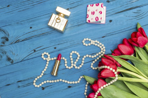Red tulips in the necklace, lipstick and bottle of perfume on the background of blue wooden board