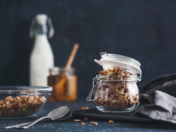 Homemade granola in glass jar on dark table. Ingredients for healthy breakfast - granola, milk and honey. Copy space for text. Low key
