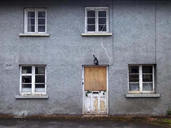 An old abandoned grey derelict house on a residential street with dirty broken windows and a boarded up door