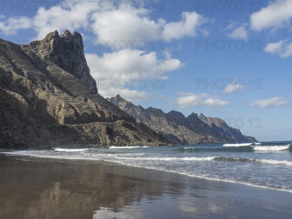 A peaceful beach with rocky cliffs and gently breaking waves under a blue sky and white clouds, lava beach with mountains and blue sky in the background, tenerife, spain