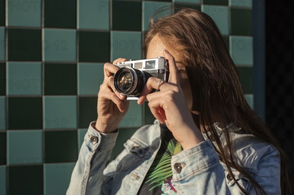 Cool girl model with retro film camera wearing a denim jacket, dark hair outdoors over city wall in a cage background. Having fun in the city with camera, travel photo of photographer