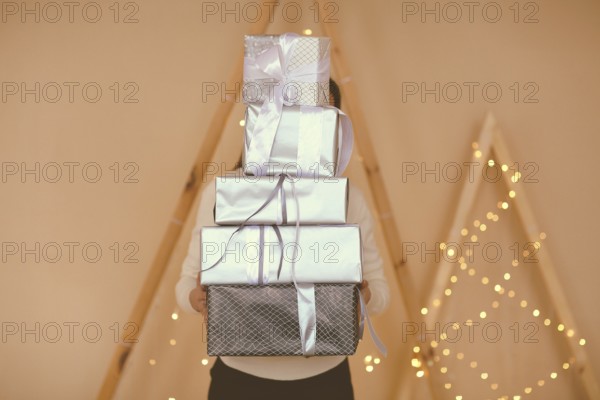 Female holding big stack of presents wrapped in brown paper with white bows on simple background with lights
