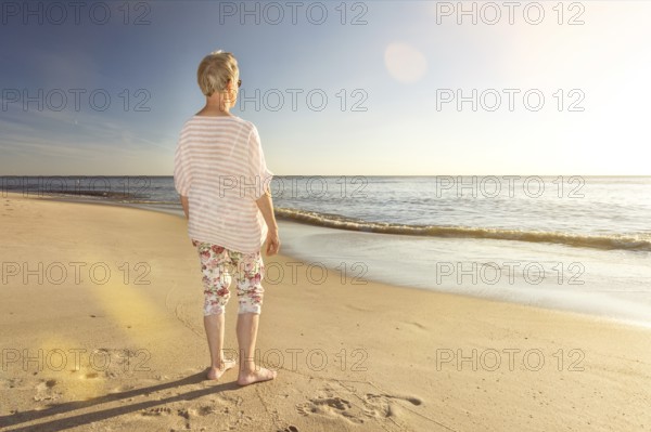 Older woman standing at the beach and looking towards the ocean with scenic lens flare