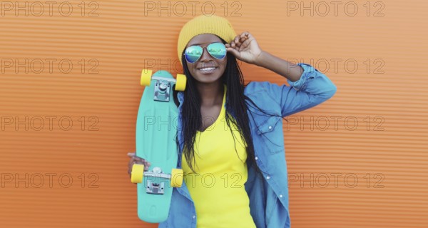 Summer portrait of stylish happy African young woman with skateboard, modern black teenage girl in yellow hat, glasses posing on orange background