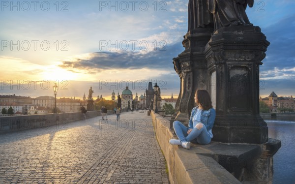 Young brunette woman, sitting relaxed on the Charles Bridge walls, over the Vltava river, admiring the sunrise over Prague City, in Czech Republic
