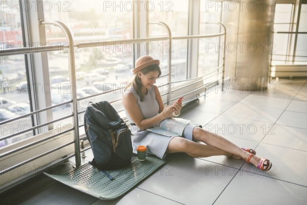 Theme of tourism and travel of young student. Beautiful young caucasian girl in dress and hat sits on floor tourist rug inside terminal airport terminal. Waiting room delayed flight, delay departure