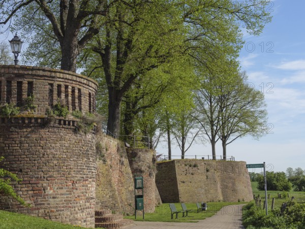 Defence defence tower and brick wall along a walkway, flanked by trees and park benches under a blue sky, beautiful promenade along the Rhine with a tower, lanterns and trees, springtime on the Rhine, Rees, Germany