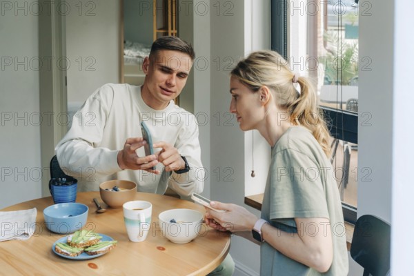 Ein Ehemann zeigt seiner Frau die Nachrichten auf seinem Handy, während er beim Frühstück am Tisch sitzt