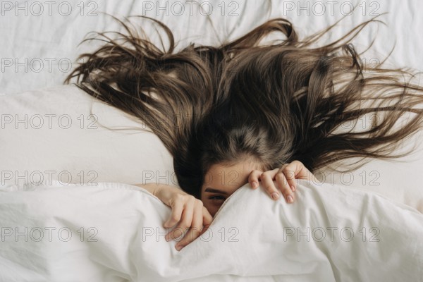 Junge lustige brünette Frau versteckt sich unter der Bettdecke und entspannt sich im Bett