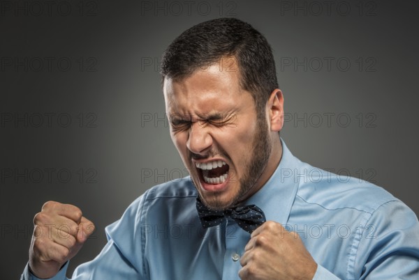 Portrait of angry upset young man in blue shirt and butterfly tie with fists up yelling isolated on gray studio background. Negative human emotion, facial expression. Closeup