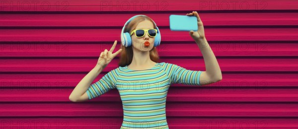 Summer portrait of stylish young woman taking selfie with smartphone listening to music with headphones, modern cool girl using phone blowing a kiss on pink background