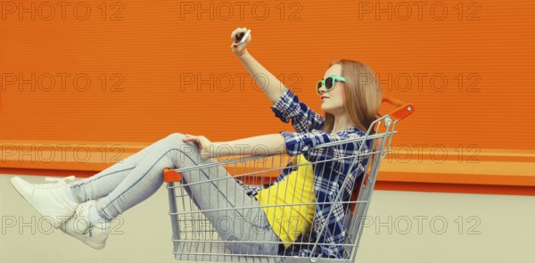 Stylish happy young woman taking selfie with phone, cool modern teenage girl having fun sitting in shopping trolley cart in the city on a summer day