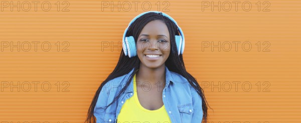 Portrait of happy African young woman listening to music with headphones, cool modern black teenage girl smiles on colorful bright orange background, summer day