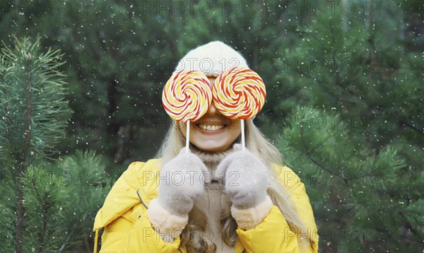 Happy funny smiling young woman having fun with lollipop enjoys snow, in winter park against Christmas tree, joyful girl standing in snowy forest with snowflakes, festive background