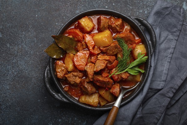 Beef meat stew with potatoes, carrot and delicious gravy in black casserole pot with bay leaves and fresh green herbs with spoon on black dark rustic concrete background from above, food photography