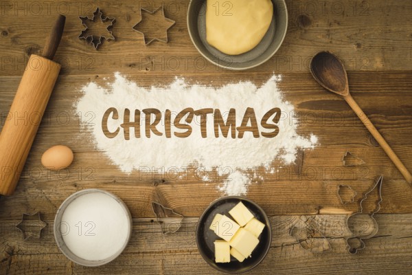 Collection of ingredients and utensils for baking Christmas biscuits with the word Christmas written in flour in the centre of the picture on a dark wooden background